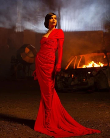 Elegant woman in a red gown stands near a burning car against a dimly lit, apocalyptic background, creating a striking contrast.