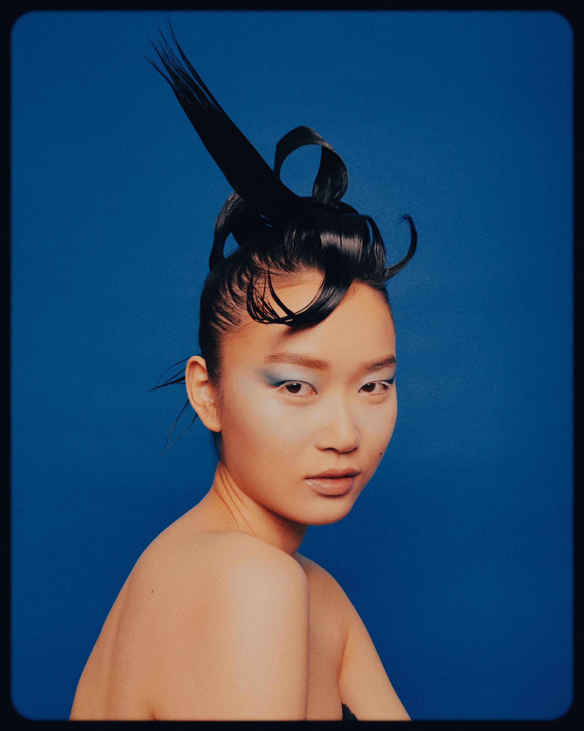 Portrait of a nude Asian female with an elaborate black hairstyle and blue eyeshadow against a solid blue backdrop.