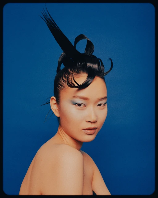 Portrait of a nude Asian female with an elaborate black hairstyle and blue eyeshadow against a solid blue backdrop.