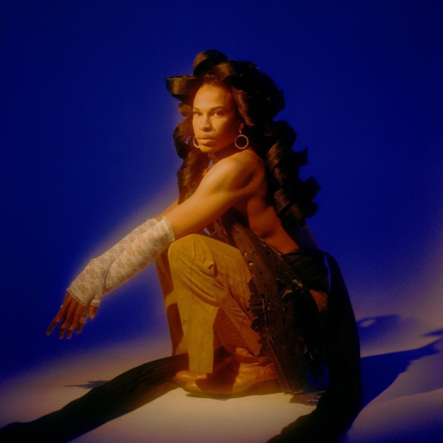Portrait of a slender adult female in tan pants and black top, accessorized with white lace arm warmers, illuminated against a deep blue backdrop.