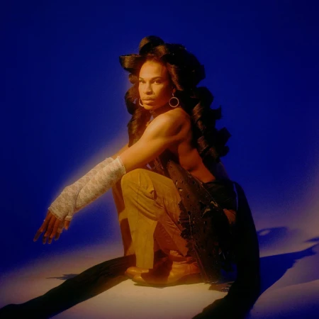 Portrait of a slender adult female in tan pants and black top, accessorized with white lace arm warmers, illuminated against a deep blue backdrop.