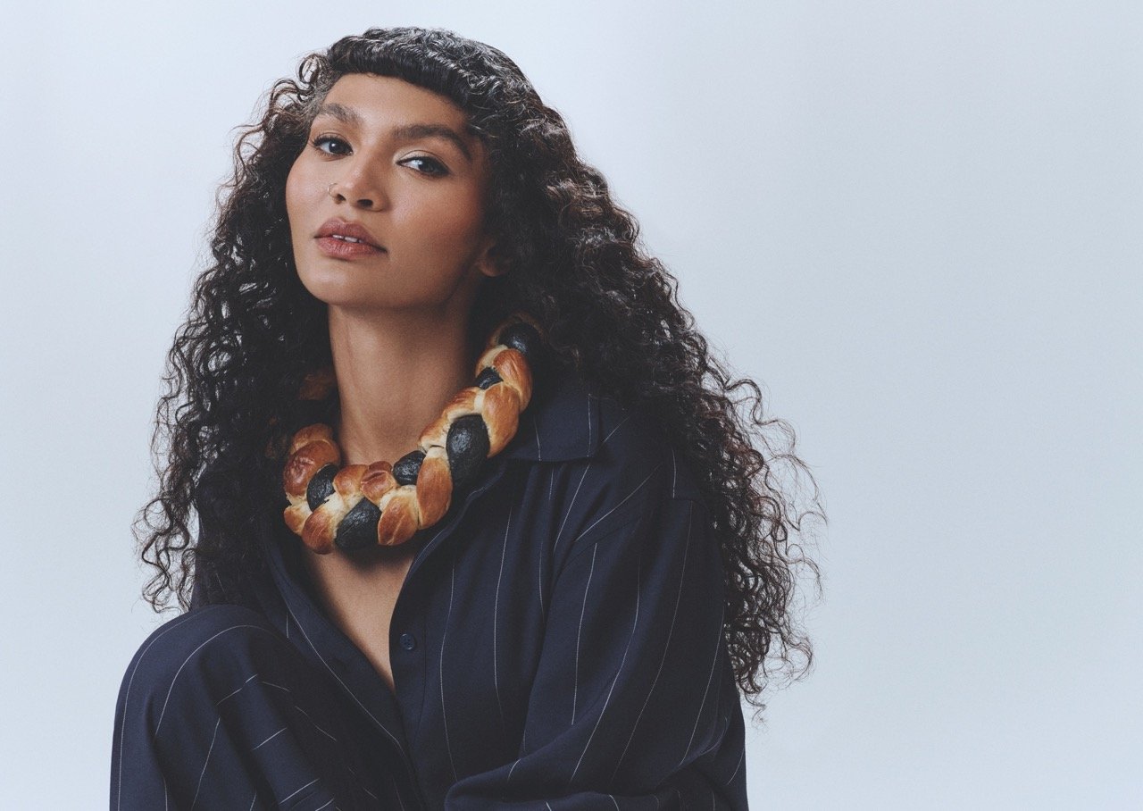 Woman with curly hair wearing a braided bread necklace and a pinstripe shirt, posing in a brightly lit photo studio.