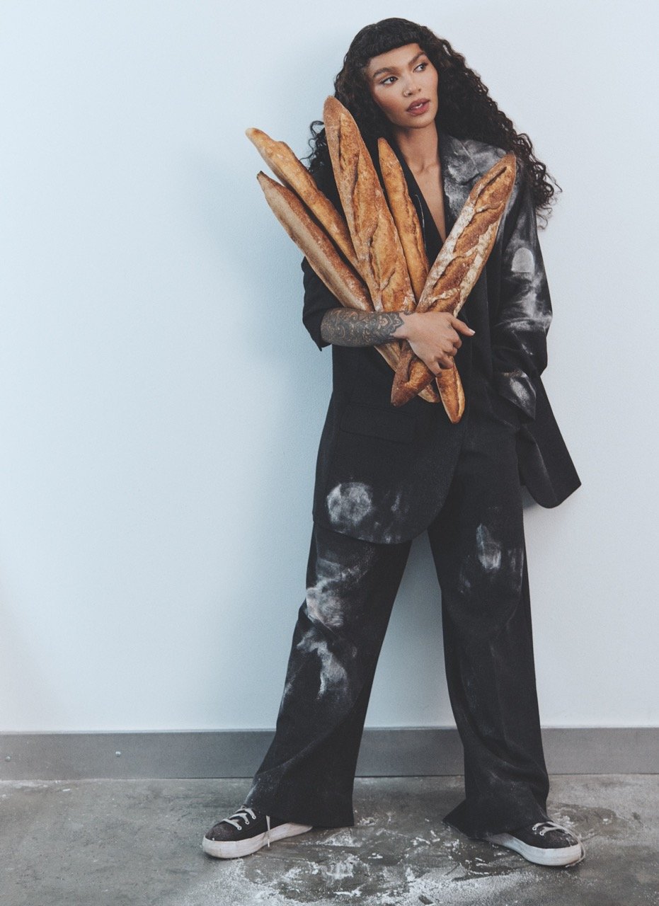 Woman in black suit holds several baguettes in a studio setting with white wall and concrete floor, showcasing fashion and food.