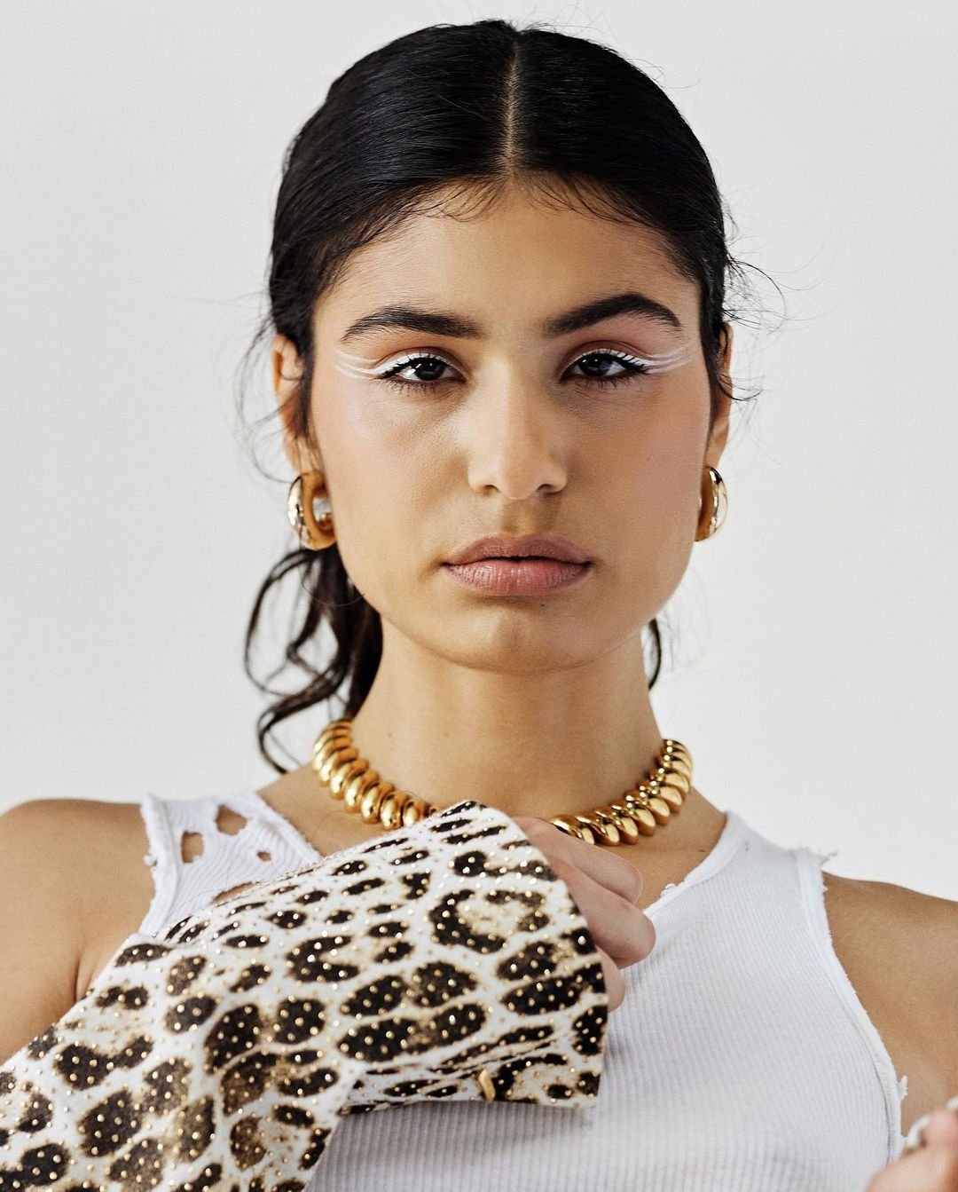 A woman with white eyeliner and a gold choker poses in a white tank top and leopard-print jacket against a neutral background, shot in portrait.