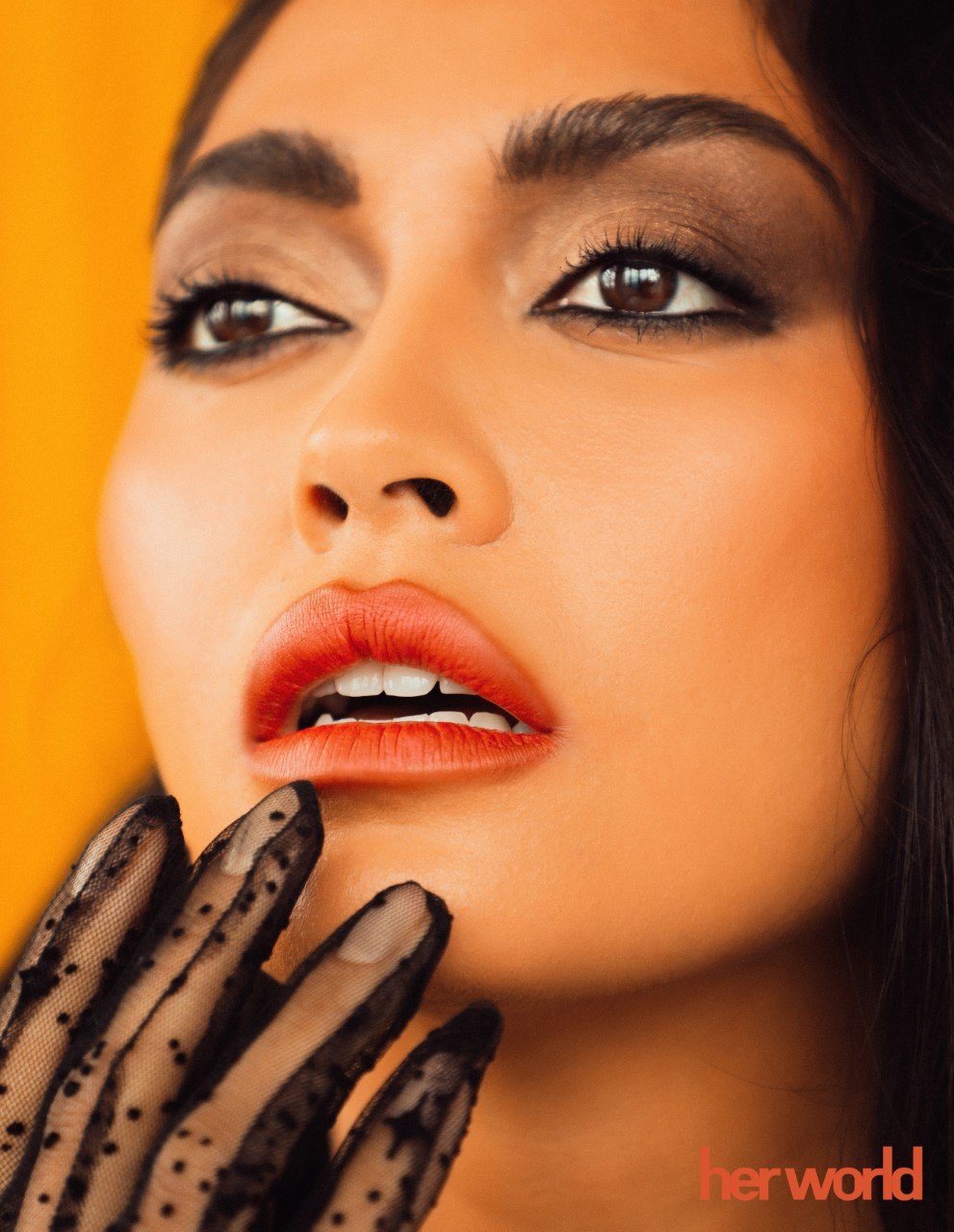 Close-up portrait of a woman with dark hair and dramatic makeup, wearing sheer polka dot gloves against a warm orange background.