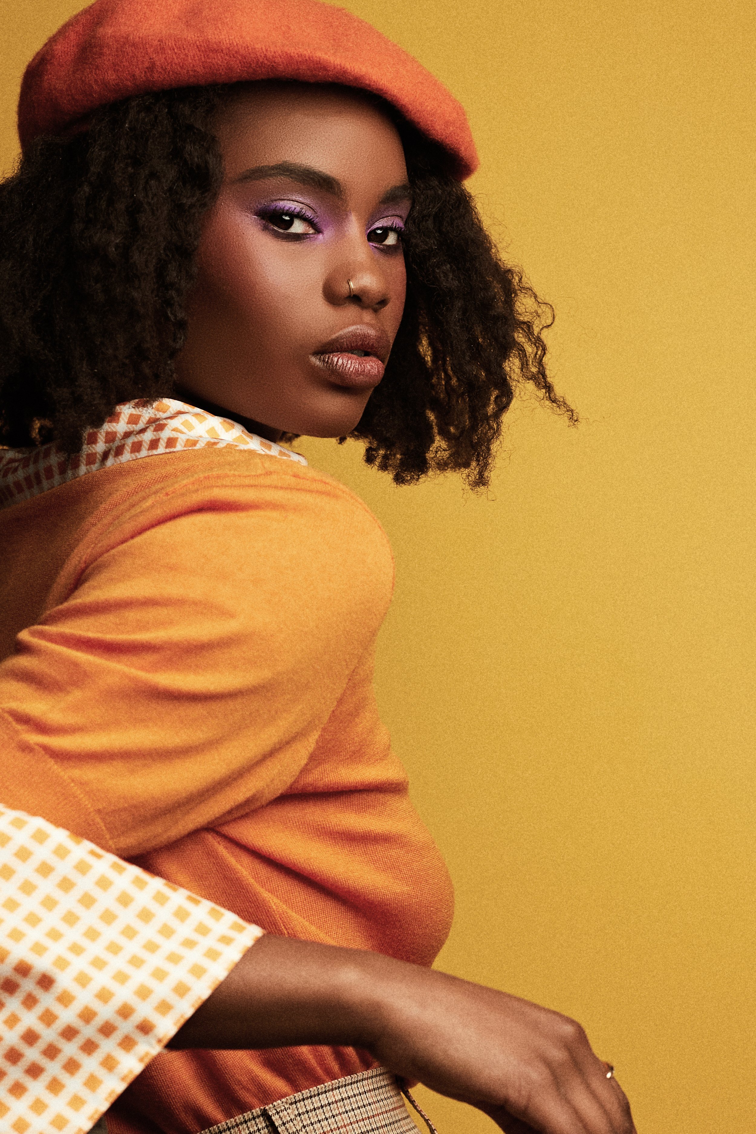 Headshot of a young Black woman wearing a red-orange beret and orange top against a yellow backdrop, with purple eyeshadow makeup.