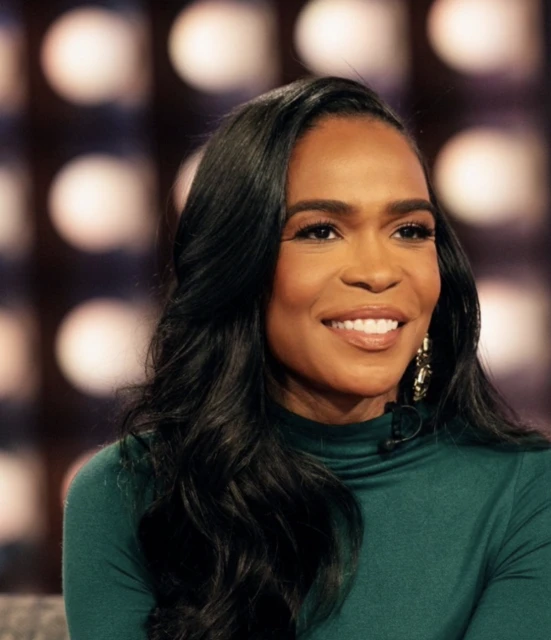Close-up shot of a smiling adult female with long black hair, wearing a green turtleneck, and gold earrings against a blurred background.