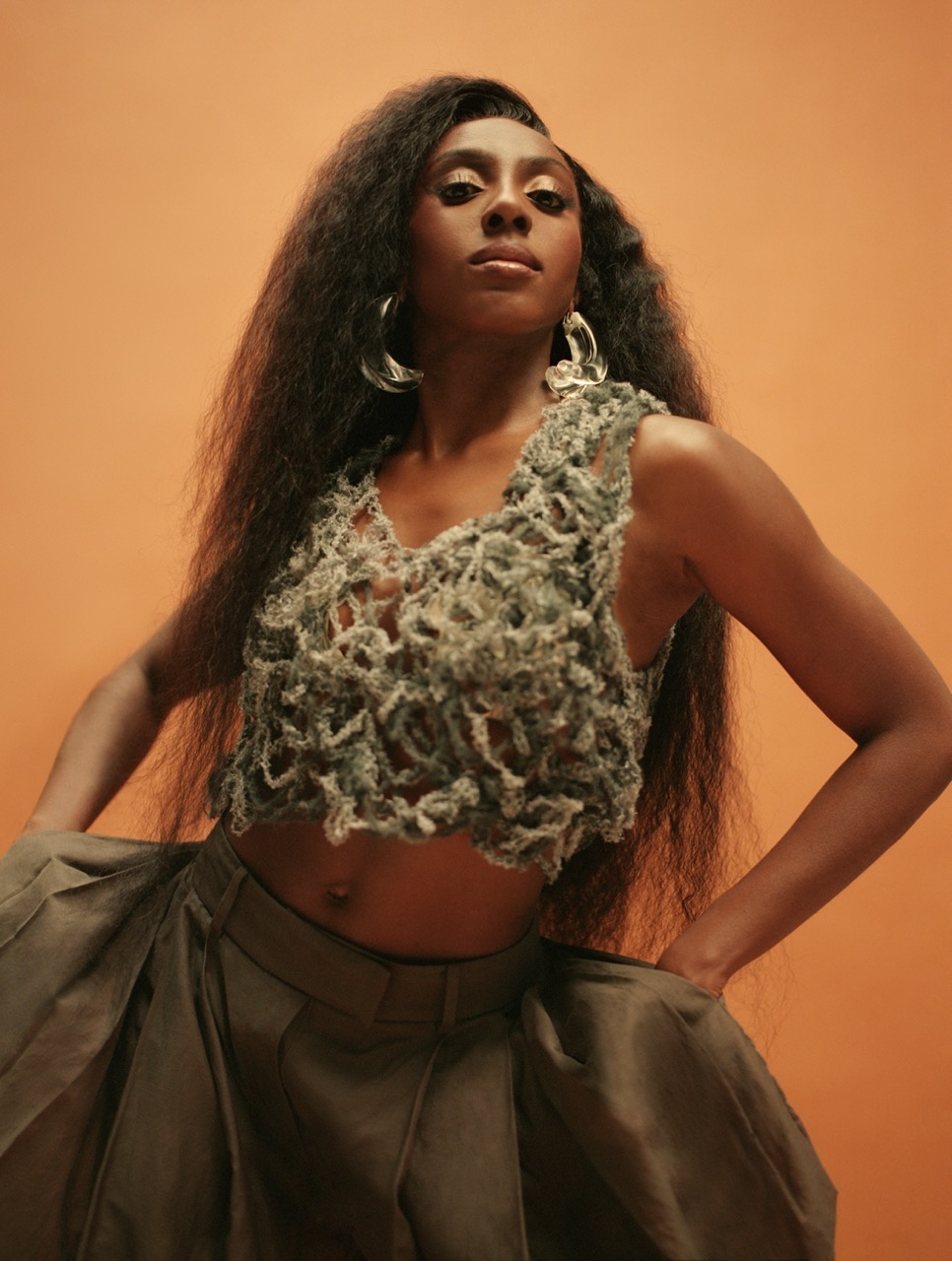 Elegant woman with long dark hair poses in a crochet top and flowing skirt against a warm orange background in a fashion editorial.
