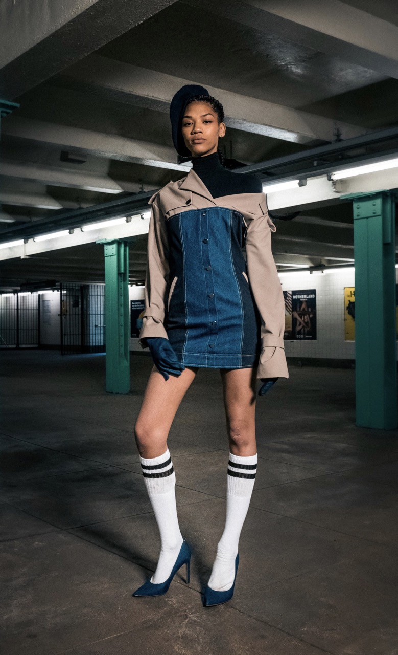 Woman in denim dress, khaki jacket, and white knee-high socks poses in subway with striped ceiling and tile walls for fashion editorial.