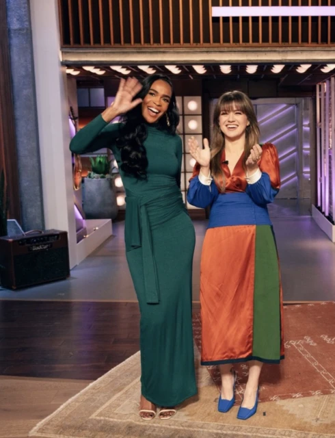 Two adult women stand in a television studio setting. One woman wears an emerald green dress and the other a colorful paneled dress.