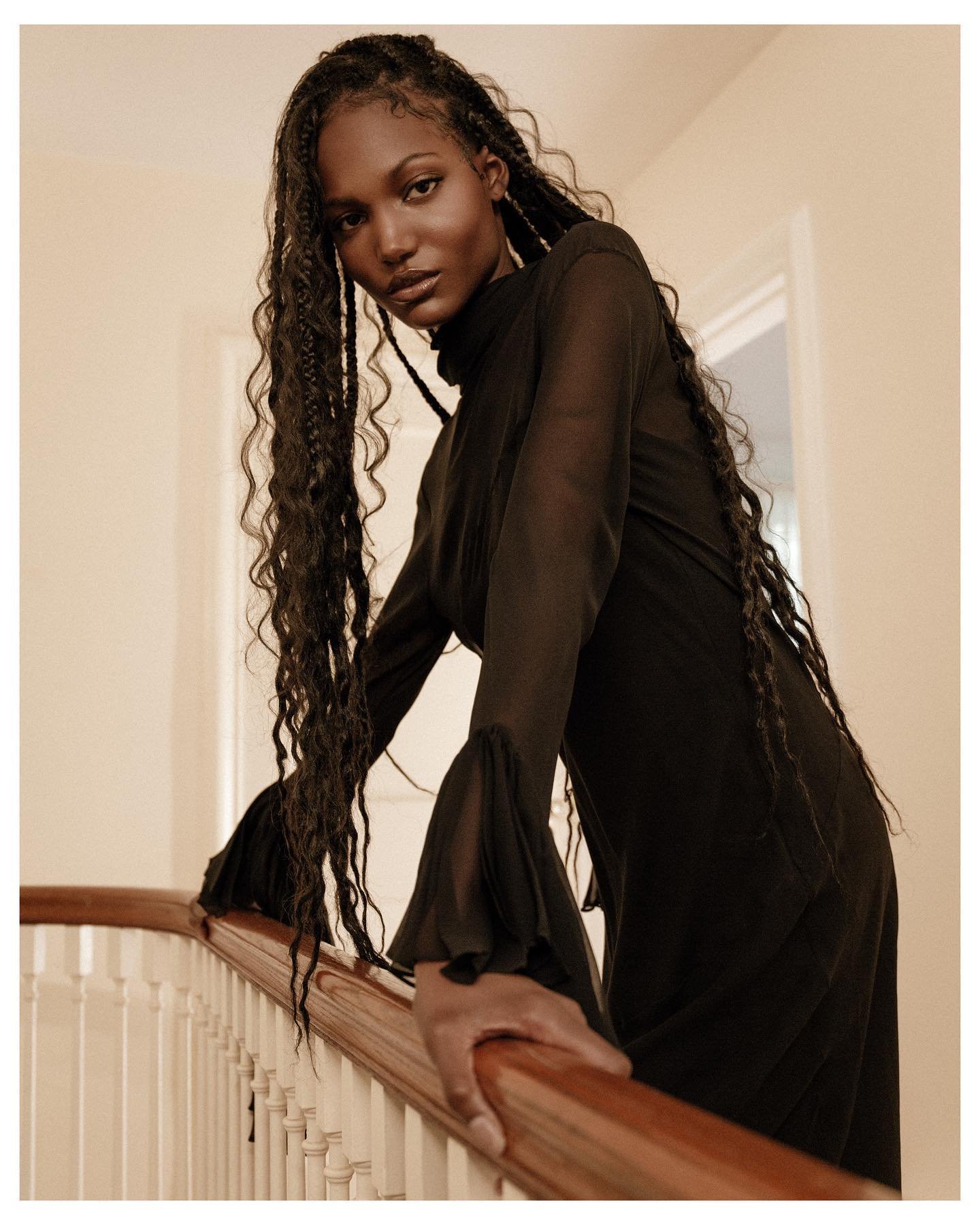Woman with long braided hair poses on stairs in a black, high-necked dress with sheer sleeves in a neutral-toned interior setting.