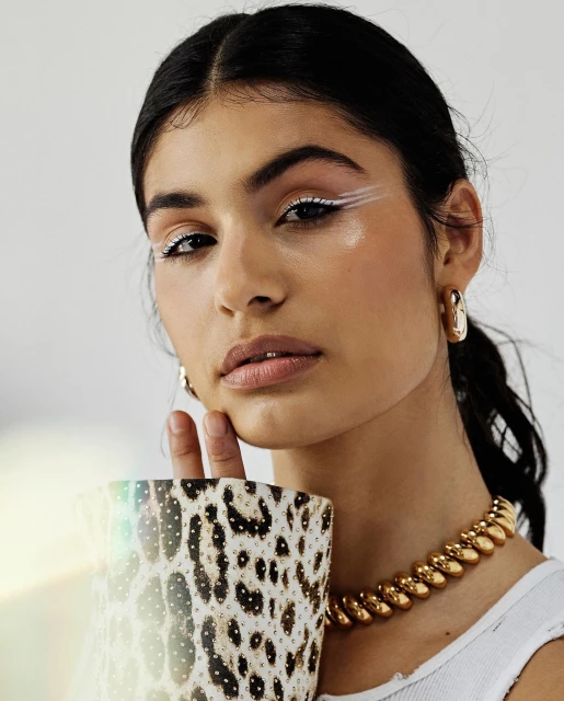A young woman with dark hair and white eyeliner poses indoors, holding a leopard-print vase and wearing a chunky gold necklace and gold earrings.