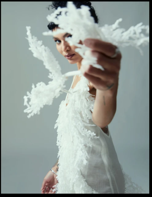 Adult female with short curly hair wears a white dress with fringed details, posing in front of a gray studio backdrop with shallow depth of field.