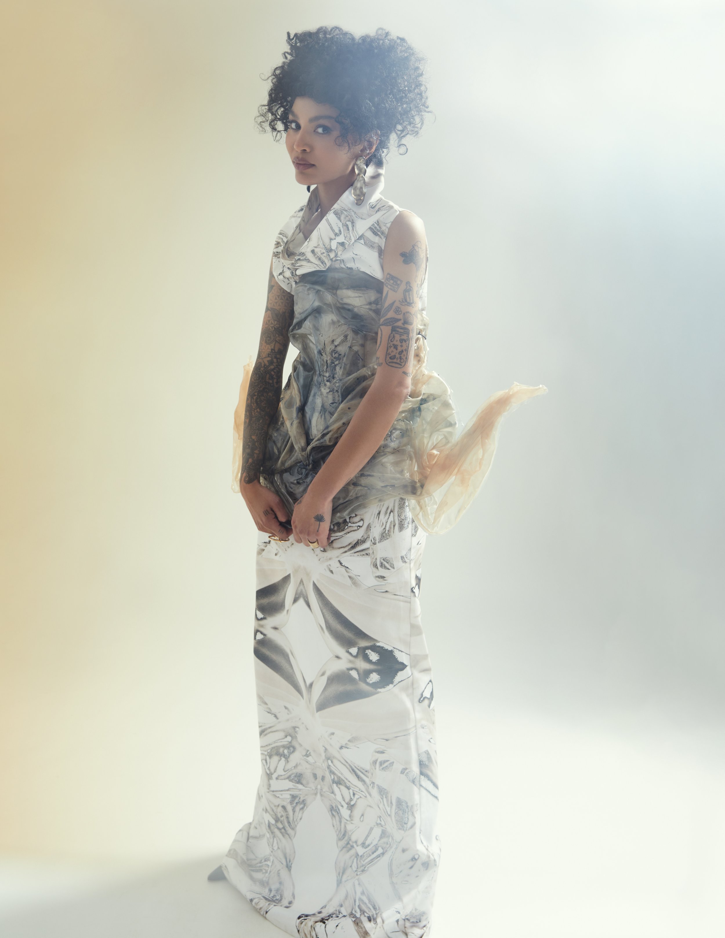A woman with curly hair poses in a sleeveless marble-patterned gown in a studio with diffused lighting and an off-white backdrop.
