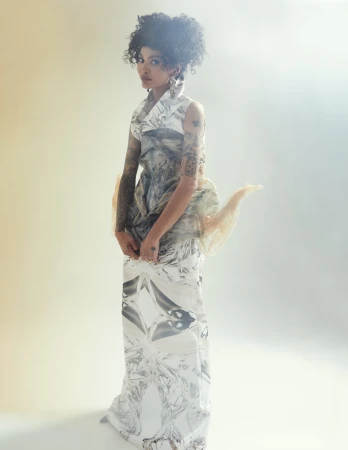 Model with curly dark hair wearing a patterned sleeveless gown against a soft white studio background with warm lighting.