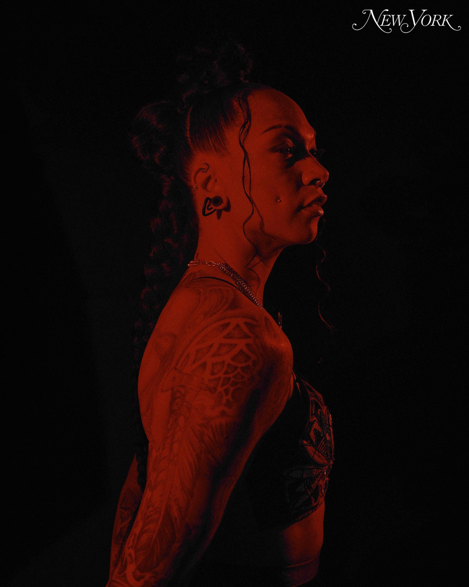 Profile shot of a tattooed Black woman with an elaborate hairstyle and jewelry, illuminated by red lighting against a dark background.