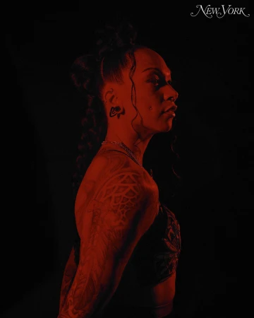 Profile shot of a tattooed Black woman with an elaborate hairstyle and jewelry, illuminated by red lighting against a dark background.
