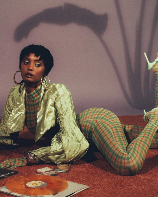 A young adult woman lying on her stomach with her legs up and vinyl records on the floor, modeling a green plaid suit.
