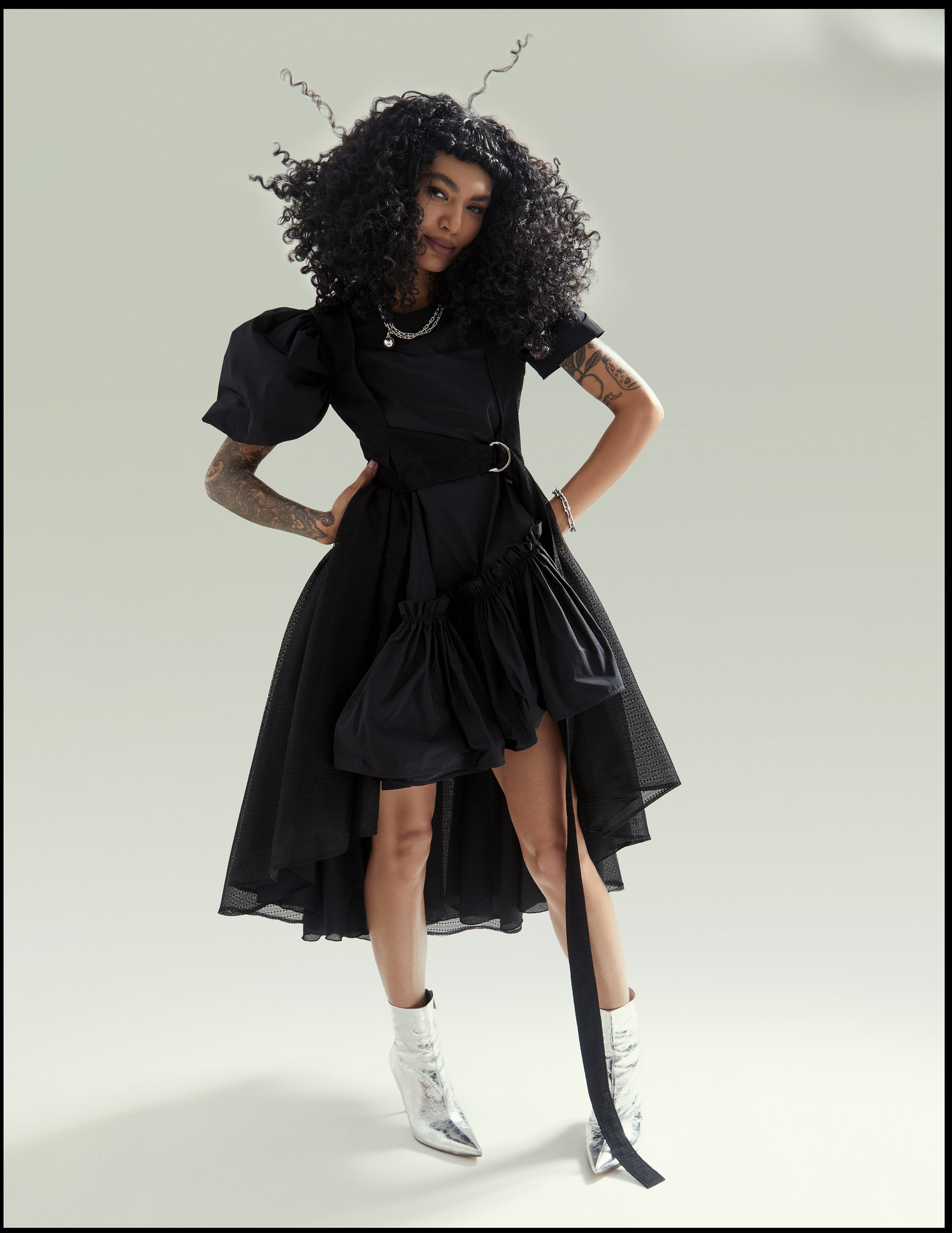 Woman in a black dress with puffed sleeves and silver boots standing against a neutral studio backdrop
