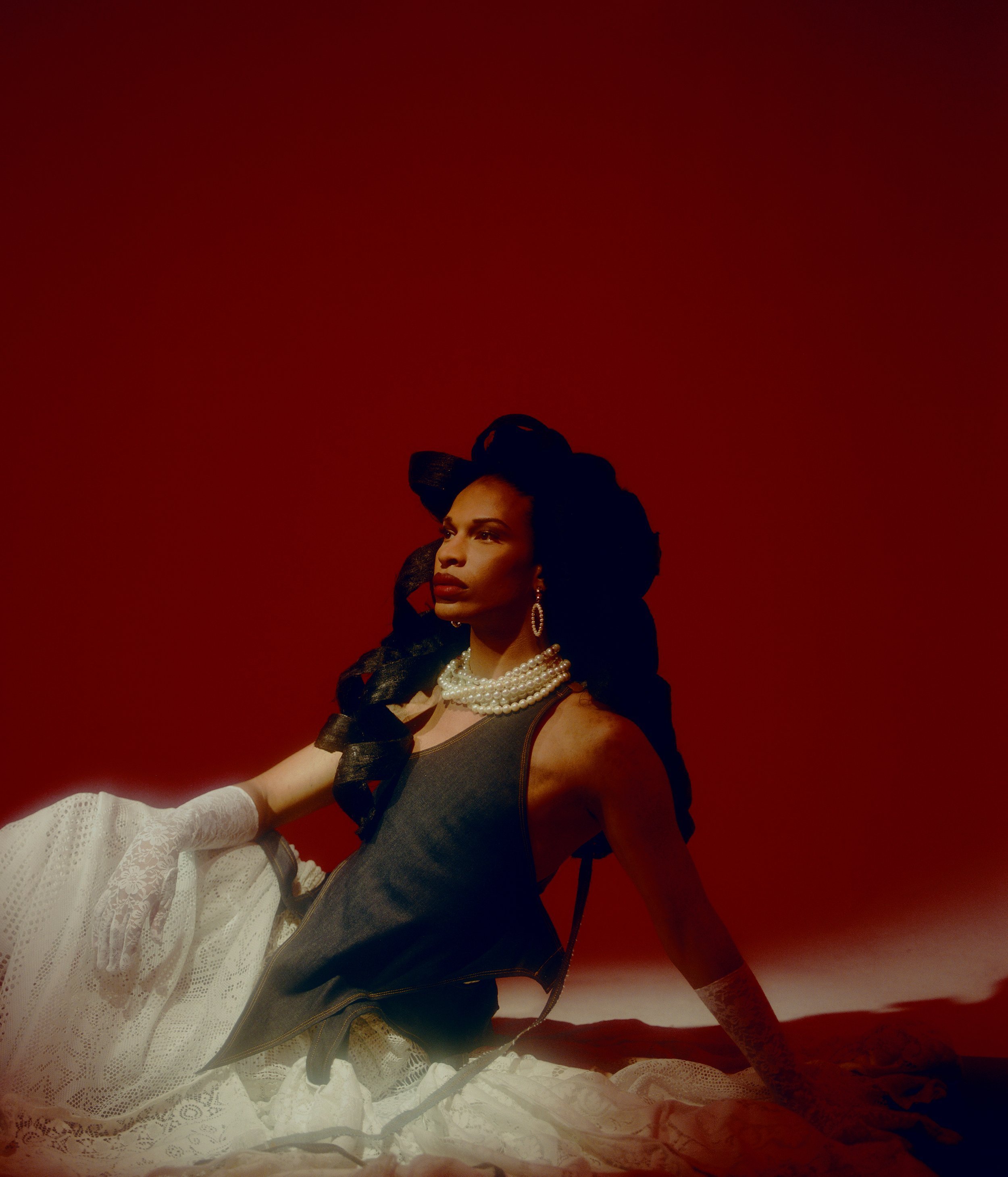 Portrait of an adult with dark hair, wearing a pearl necklace, black tank top, and lace skirt against a solid red backdrop.