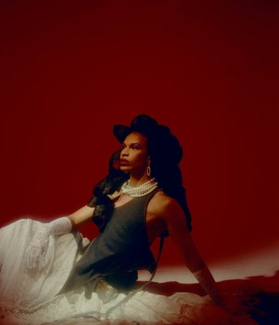 Portrait of an adult with dark hair, wearing a pearl necklace, black tank top, and lace skirt against a solid red backdrop.
