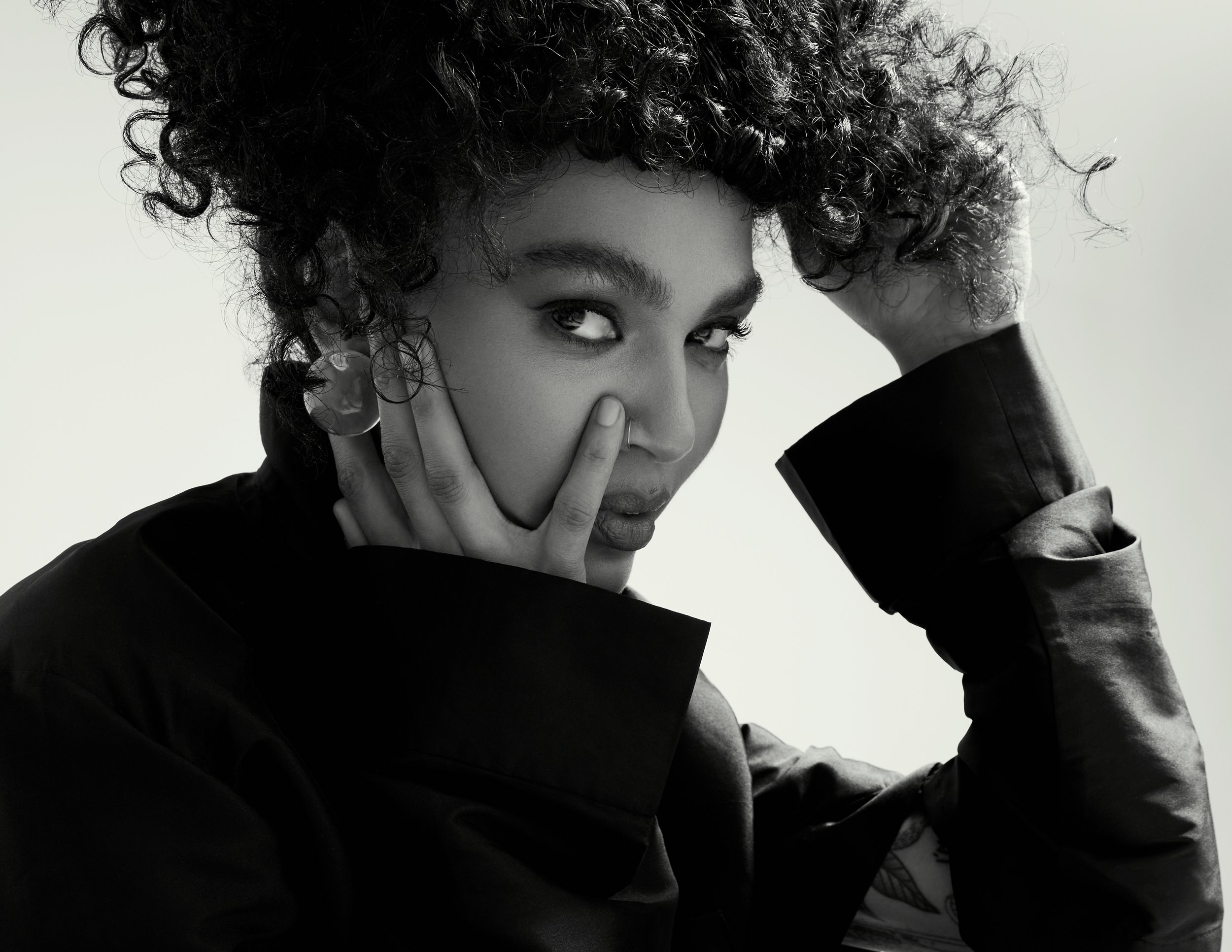 Black and white portrait of a woman with curly hair, wearing a dark top with hand near face, looking at camera.