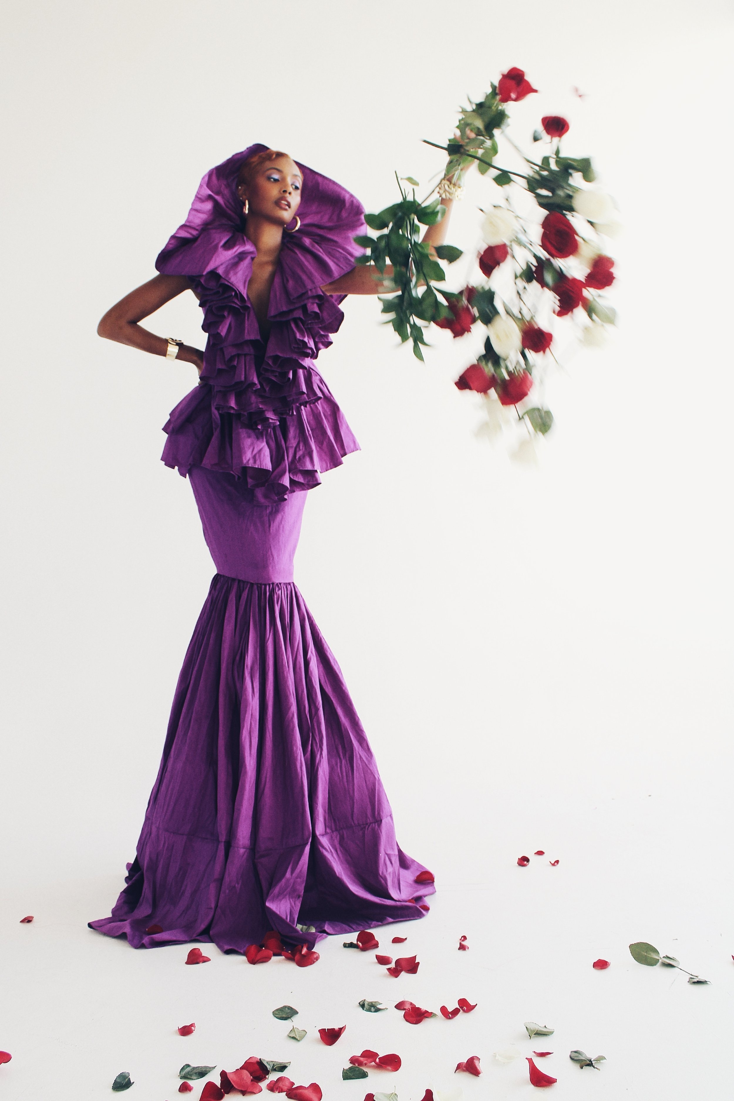 An African-American woman poses in a purple couture gown with a large bouquet of red and white roses in a fashion studio.