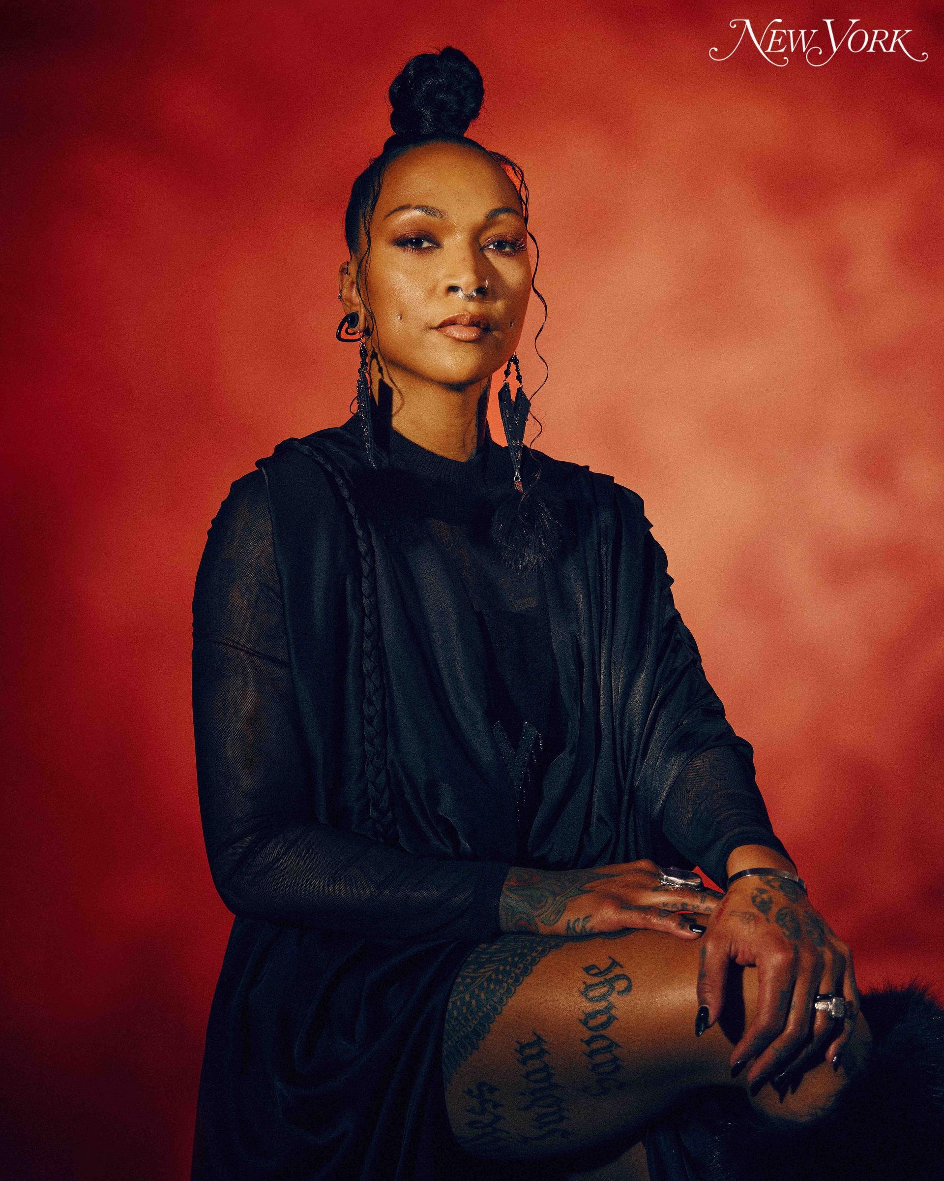 Portrait of a woman with a high bun hairstyle wearing a dark outfit in front of a red background, featuring body tattoos and dark hanging earrings.