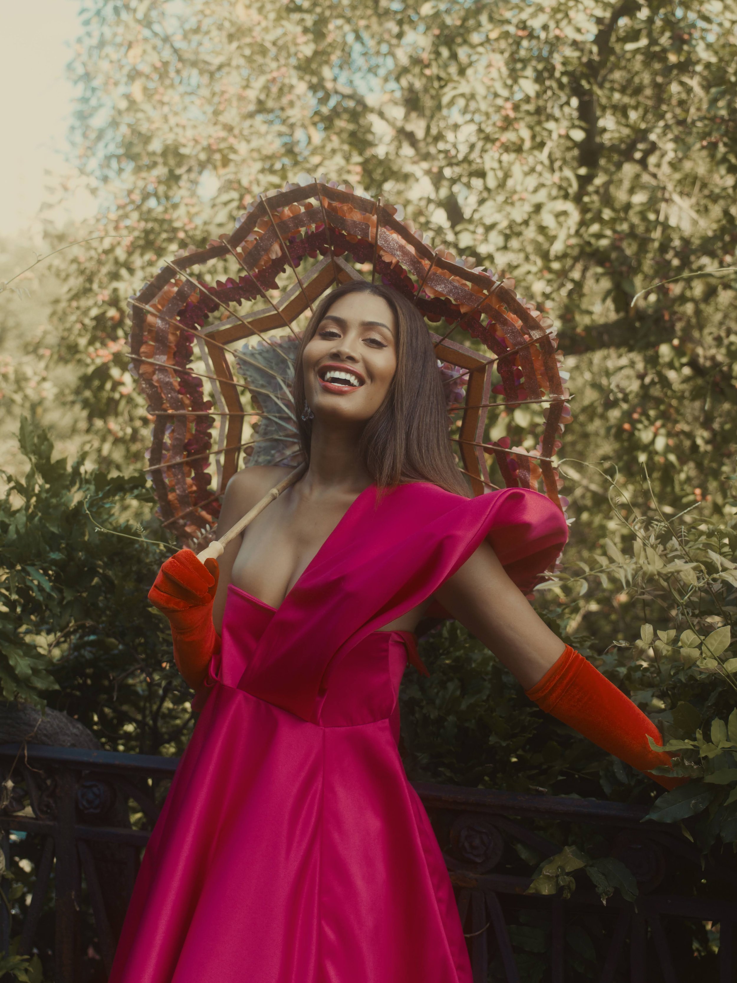 A smiling adult woman poses outdoors in a vibrant pink formal gown and orange gloves, holding a decorative parasol, with trees in the background.
