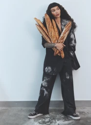 Woman with long dark curly hair wearing a black suit and holding several baguettes against a plain white background