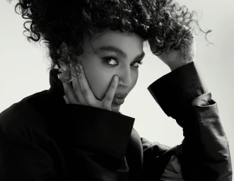 Close-up portrait of a woman with curly hair wearing a dark shirt, hand near her face with makeup by Nicole Bueno.