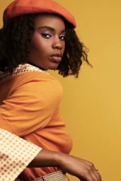 Close-up portrait of a Black woman with purple eyeshadow wearing an orange beret and patterned orange top against a yellow background.