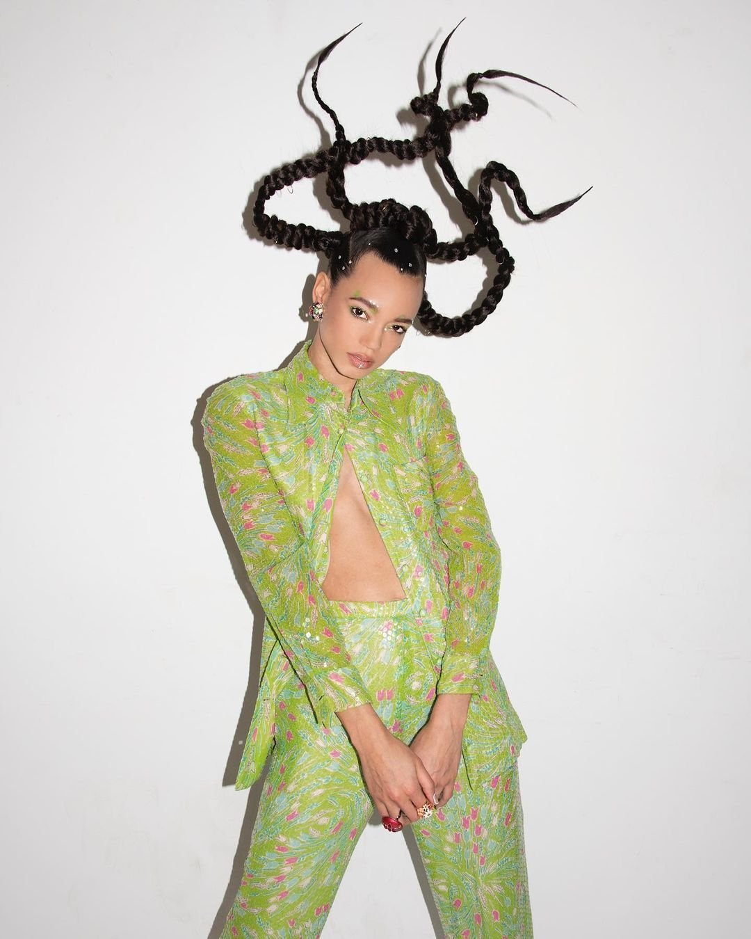 Woman in patterned green suit poses against white backdrop, showcasing striking hair design, open jacket, and confident gaze in studio setting.