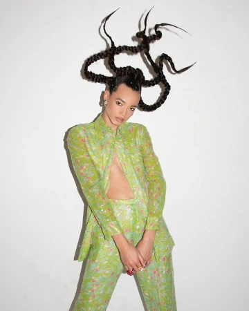 Woman in patterned green suit poses against white backdrop, showcasing striking hair design, open jacket, and confident gaze in studio setting.