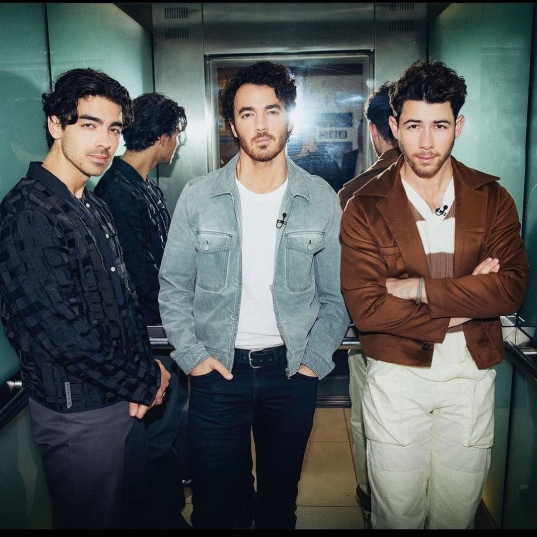 Three adult men stand in an elevator, posing in jackets and casual pants. The men are reflected in a mirror. Elevator background.
