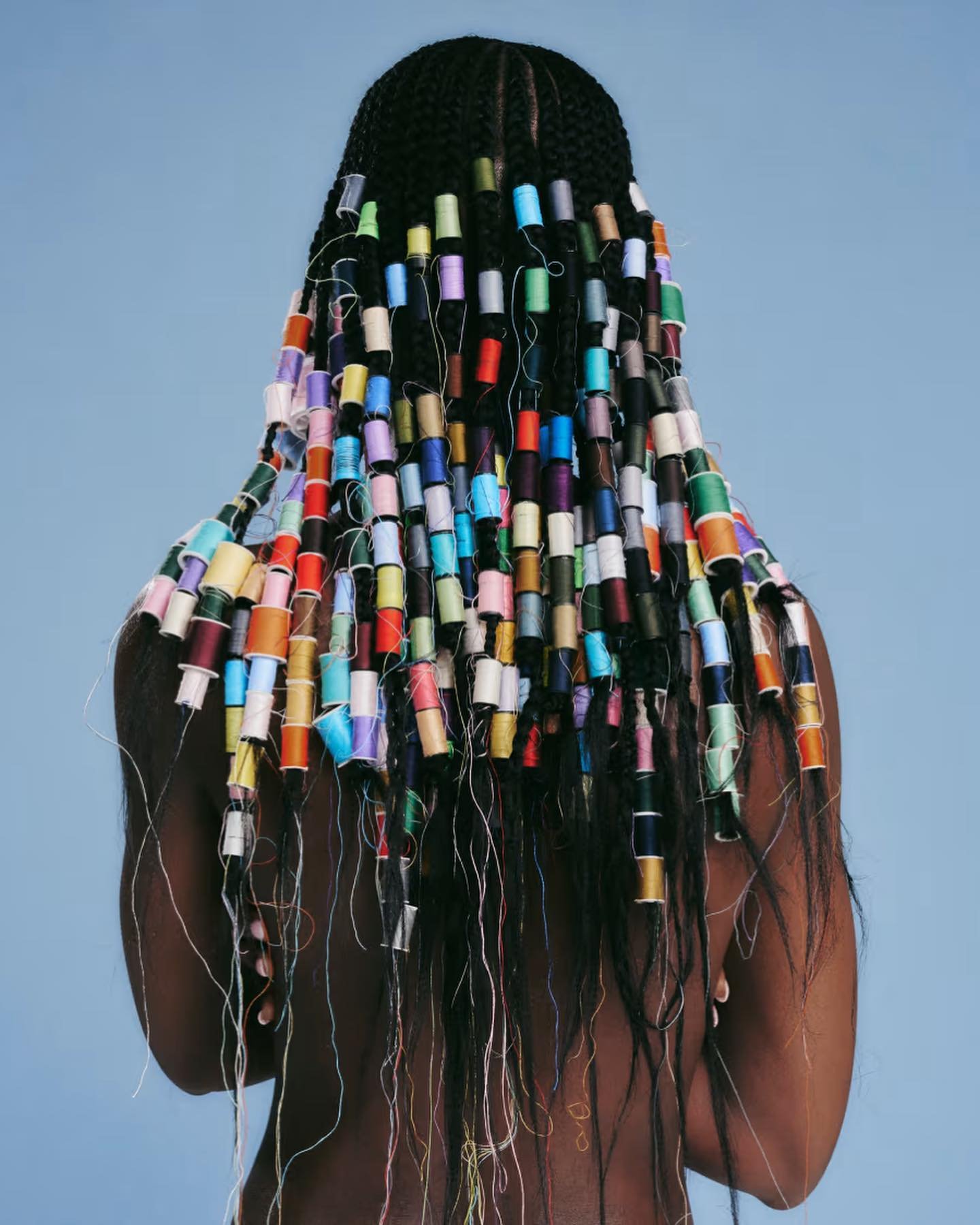 An adult woman is seen from the back, with black braided hair decorated with colorful spools of thread against a light blue background.
