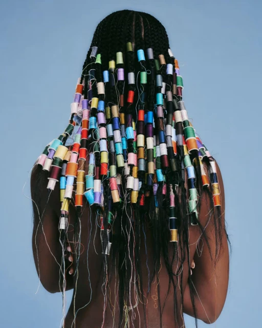An adult woman is seen from the back, with black braided hair decorated with colorful spools of thread against a light blue background.
