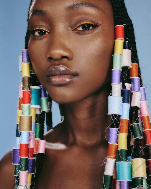 Close-up portrait of an African American female model with colorful thread spools in her braided hair against a light blue background.