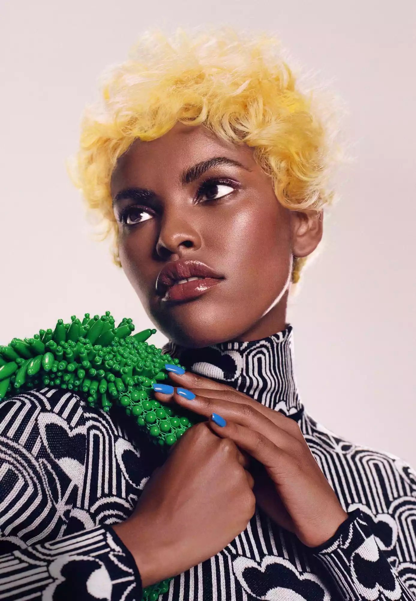A close-up studio shot of a beautiful Black woman with short, curly blonde hair, wearing a patterned top and holding a green beaded object.