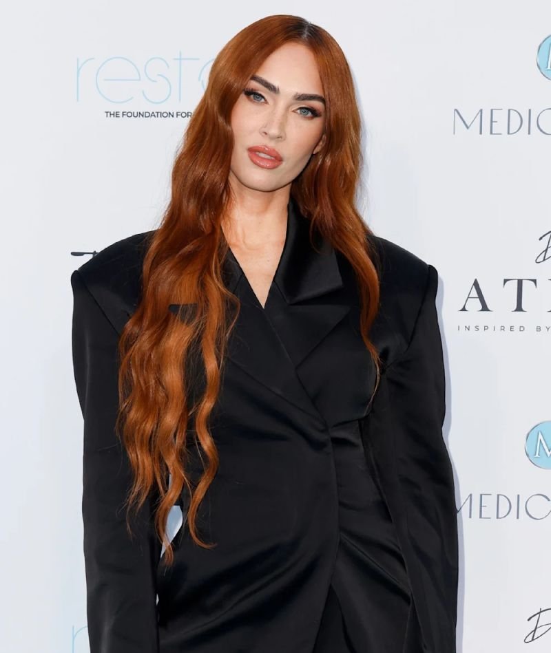 Megan Fox poses in a black blazer dress at an event with bright lighting. She has long red hair and is wearing makeup.