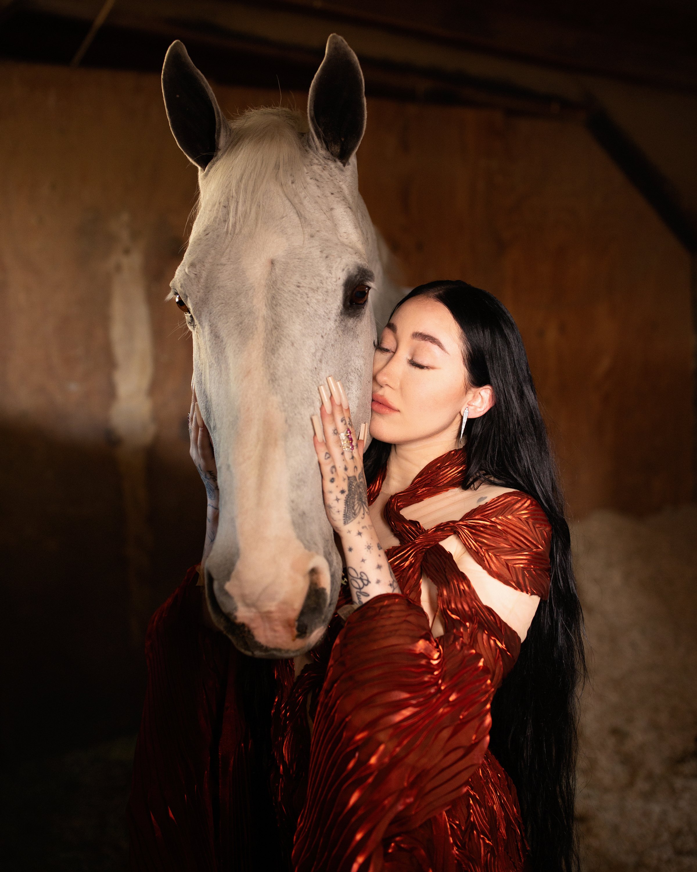 Woman embraces white horse indoors. She wears a copper dress and has long black hair. Low-key natural lighting emphasizes a tender moment.