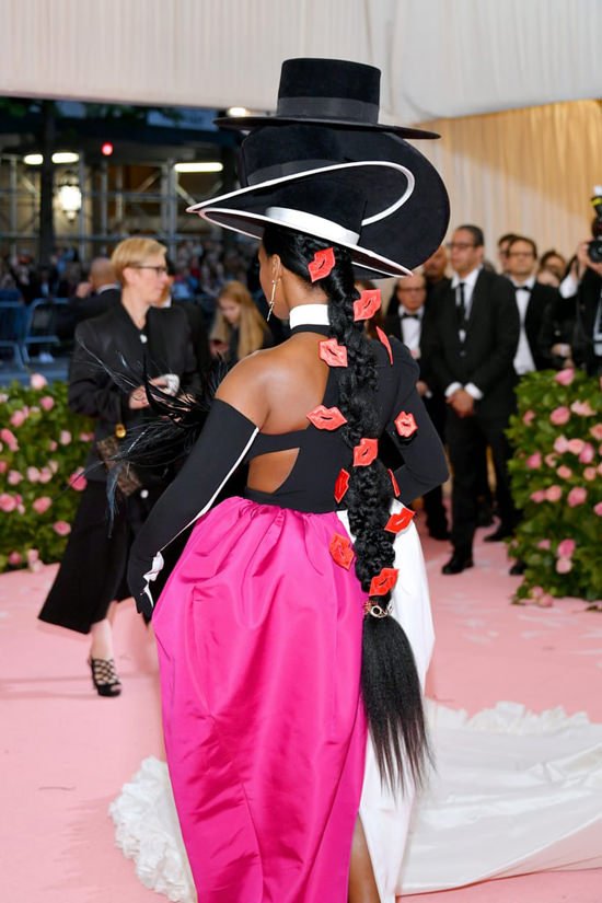 Woman in haute couture with large hat and pink dress standing on pink carpet surrounded by crowd at the Met Gala event.