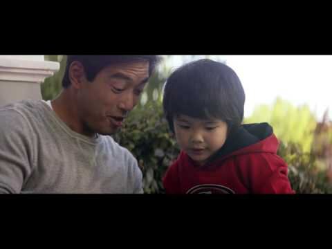 Asian father and child are outdoors together. The father wears a grey shirt and the child wears a red hooded sweatshirt.