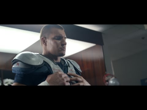 American football player fastens his shoulder pads in a locker room, preparing for a game, captured with medium shot and indoor setting.