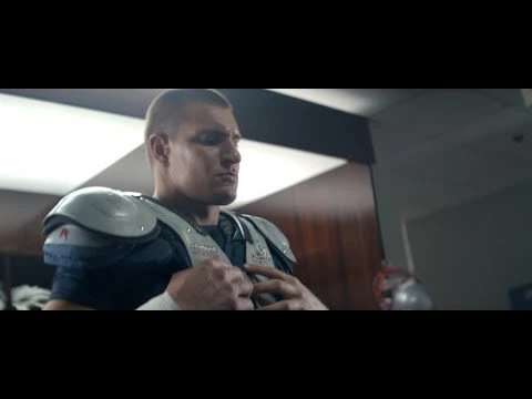 American football player fastens his shoulder pads in a locker room, preparing for a game, captured with medium shot and indoor setting.