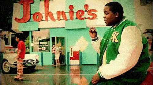 Scene features an adult male in a green letterman jacket outside Johnie's Coffee Shop with a classic car and other people visible.