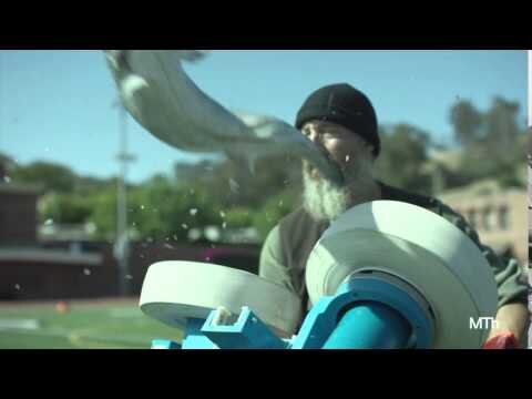 Adult male with a white beard is operating a device for hurling sports balls, set on a sports field with buildings in the background.
