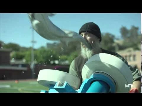 Adult male with a white beard is operating a device for hurling sports balls, set on a sports field with buildings in the background.