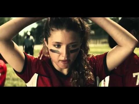 Teenage female American football player with eye black, adjusting her ponytail on an outdoor field during a bright, sunny day.