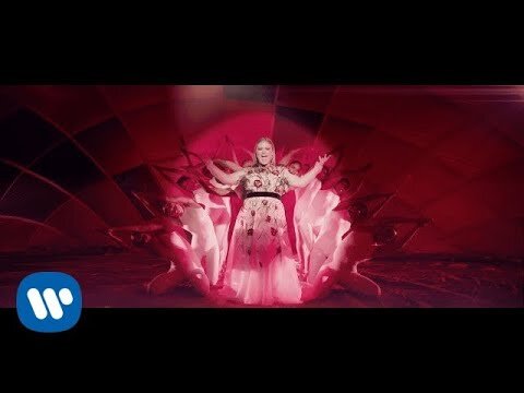 Music video scene featuring a singer in a floral gown surrounded by dancers against a backdrop of red and geometric patterns.